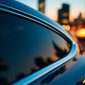 Close-up of a sleek black car's driver-side window, showcasing professional window tinting in Houston with a subtle dark tint. The sun glints off the glass, reflecting a blurred Houston street scene with palm trees, taken with a Hasselblad X2D 100C.
